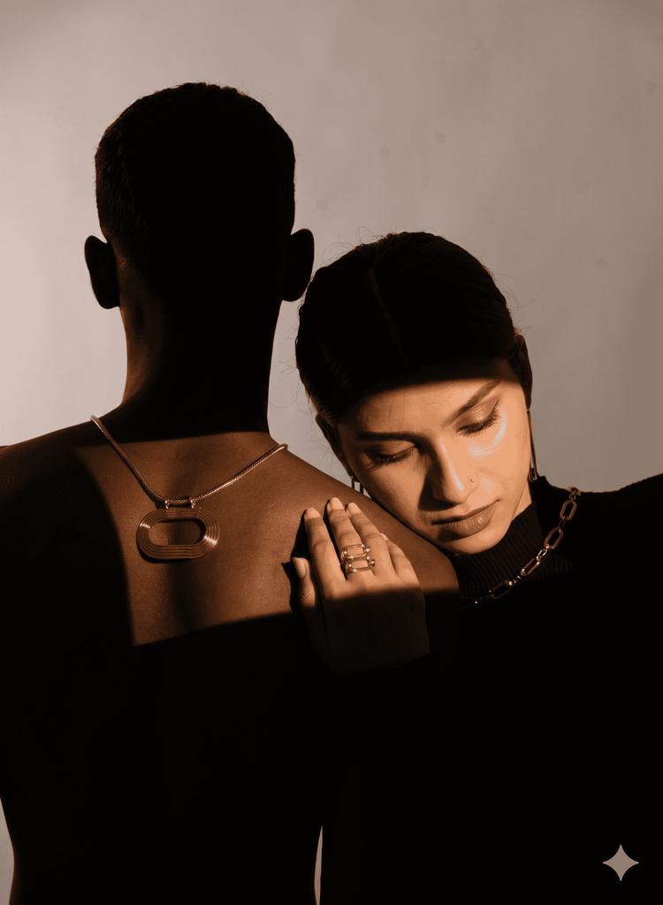 A moody fashion photograph featuring a model from behind wearing a large geometric silver pendant necklace, with another model resting their head on their shoulder wearing a gold chain necklace. Shot in dramatic, high-contrast warm sepia lighting against a neutral background.